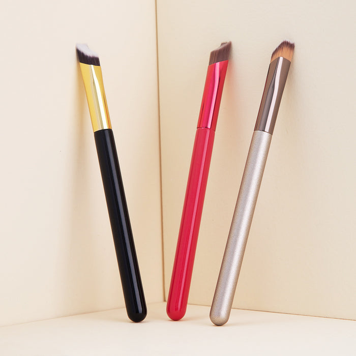 Three angled eyebrow brush for concealer with black, red, and gold handles standing against a beige background